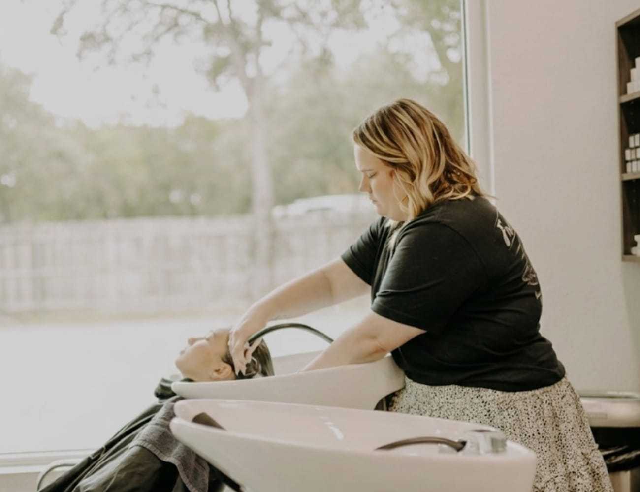 Hair stylist washing client's hair in a salon with large window view.