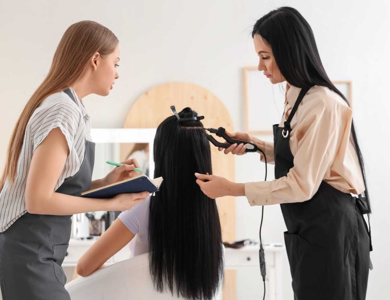 Two hairstylists work on a client's long hair using a curling iron in a salon.