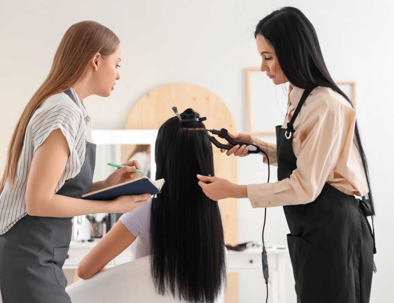 Two hairstylists work on a client's long hair using a curling iron in a salon.
