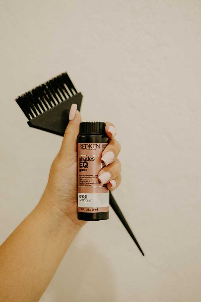 Hand holding hair dye bottle and brush against a neutral background.