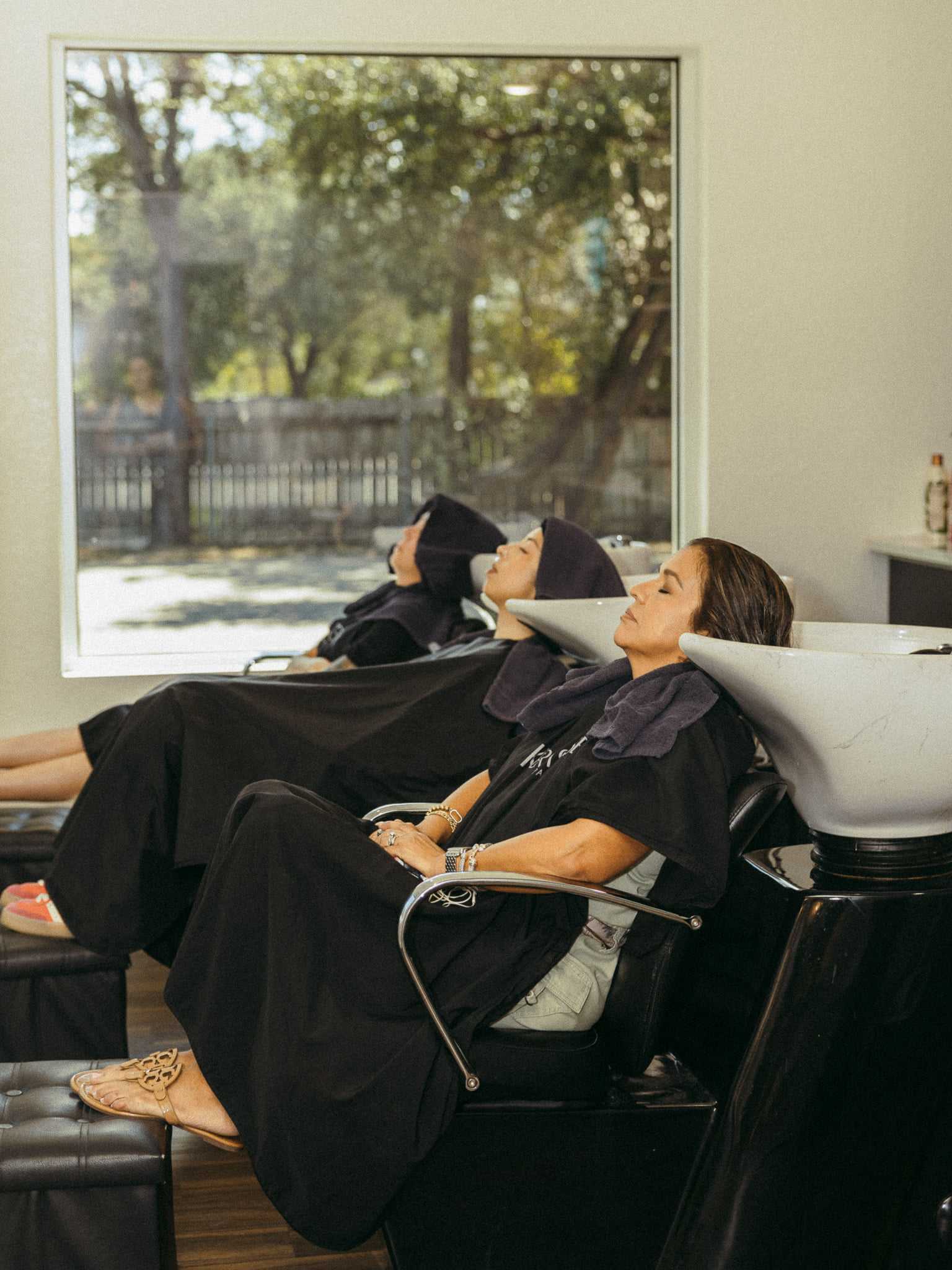 Women relax with hair towels in salon chairs, seated at wash stations near a large window.